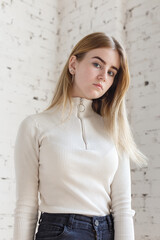 portrait of cute thoughtful teen model wearing white sweater and black jeans. caucasian skinny young female with blond hair stands in front of white brick wall. natural pretty lady standing at studio
