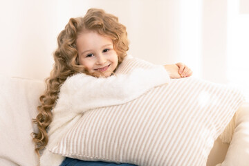 Obraz na płótnie Canvas Cute little girl with long blond hair hugging a pillow and smiling, looking into the camera, in beige interior. Happy child at home