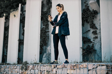 A sportswoman standing in urban exterior, taking a break and using phone.