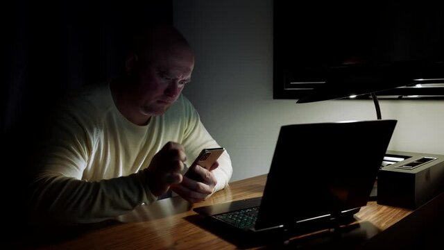 A bald, desperate middle-aged man works at a computer table on which there is a laptop, holds a smartphone in his hands and looks thoughtfully at it.