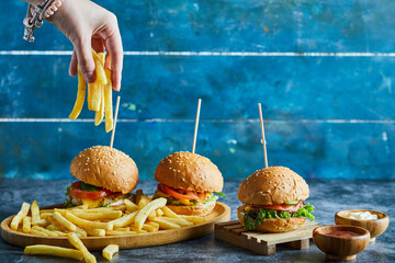A woman hand holding fry potato with three cheeseburgers, ketchup, mayonnaise on wooden plate