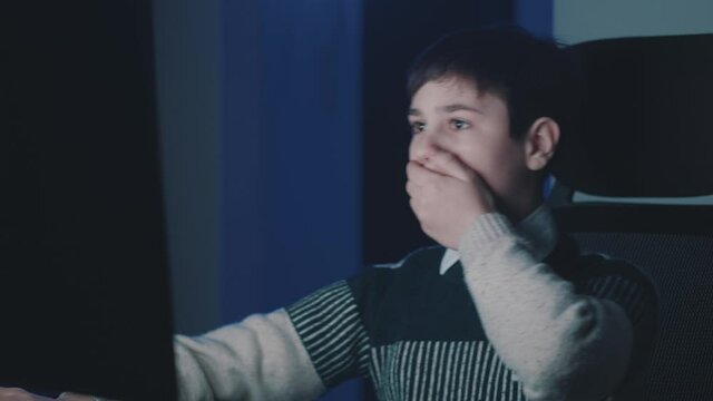 Afraid and shocked boy look at monitor screen, cover mouth with his hand while using computer laptop late at night. Schoolboy kid scared of bad news, read terrible message. Watching horror movie film.