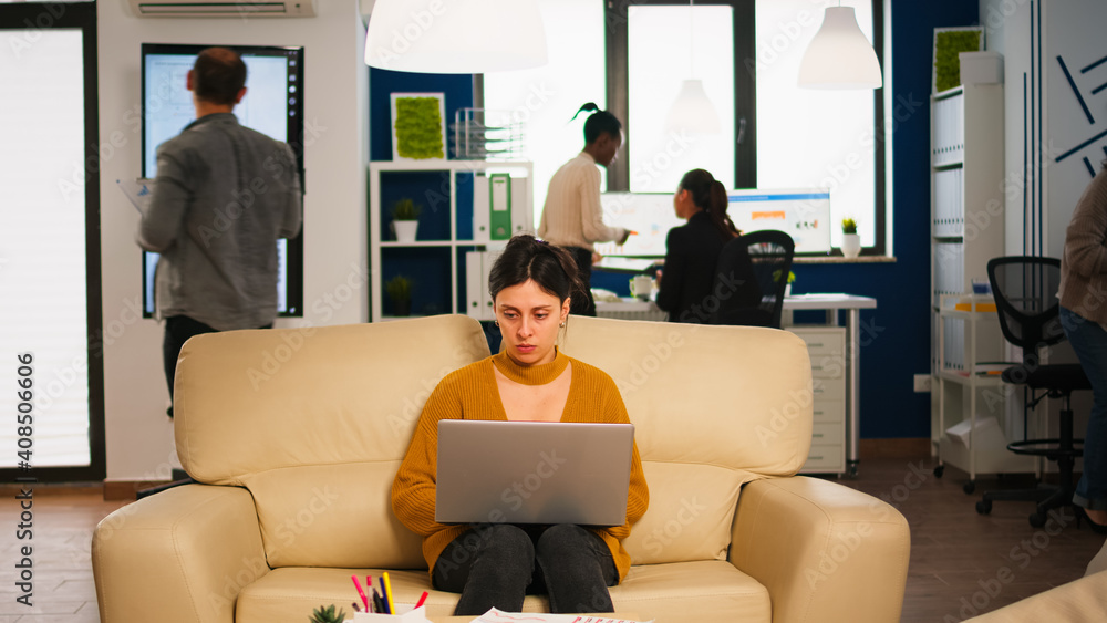 Poster concentrated manager reading mails typing on laptop sitting on couch in busy start up office while d