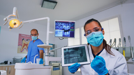 Dentist showing on tablet teeth x-ray reviewing it with patient. Doctor and nurse working together in modern stomatological clinic, explaining to old woman radiography of tooth using notebook display