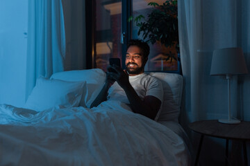 technology, internet, communication and people concept - young indian man with smartphone lying in bed at home at night