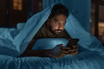 technology, internet, communication and people concept - young indian man with smartphone lying in bed under blanket at home at night