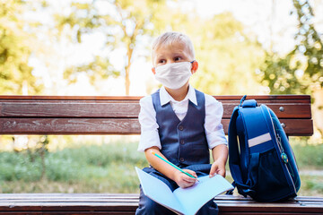 Elementary school student sits on a bench. Dressed in smart school uniform. Holding notebook on his knees and writing in it. New normal. Wears a medical protective mask for coronavirus. Covid-19