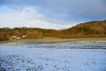 Winter landscape. A snow-covered field with green grass. Mountains covered with trees. A small village in the distance. Blue sky with clouds.