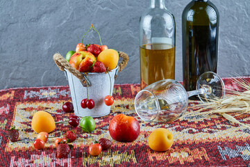 Bucket of fresh summer fruits, two bottles of wine and empty glass on carved rug