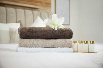 Stack of clean towels, flower and shampoo bottles on bed indoors