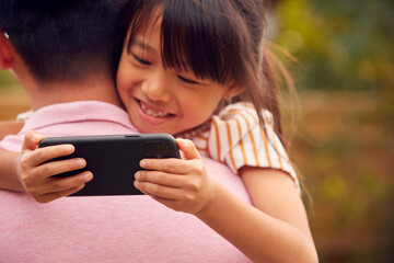 Asian Father Cuddling Daughter In Garden As Girl Looks Over His Shoulder At Mobile Phone