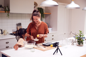 Asian Mother And Daughter Baking Cupcakes In Kitchen At Home Whilst On Vlogging On Mobile Phone