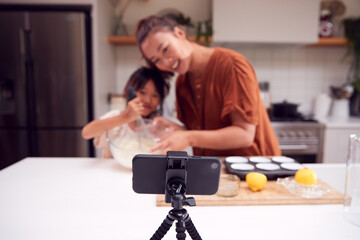 Asian Mother And Daughter Baking Cupcakes In Kitchen At Home Whilst On Vlogging On Mobile Phone