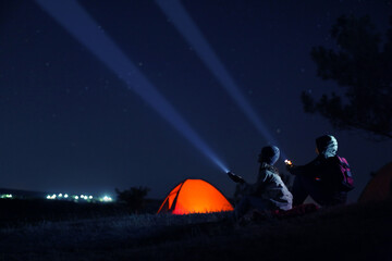 Couple with flashlights near camping tent outdoors at night - obrazy, fototapety, plakaty