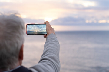 Senior man standing in front of the sea while uses his mobile phone to photograph the sunset on a winter day. Retired person enjoying retirement and freedom