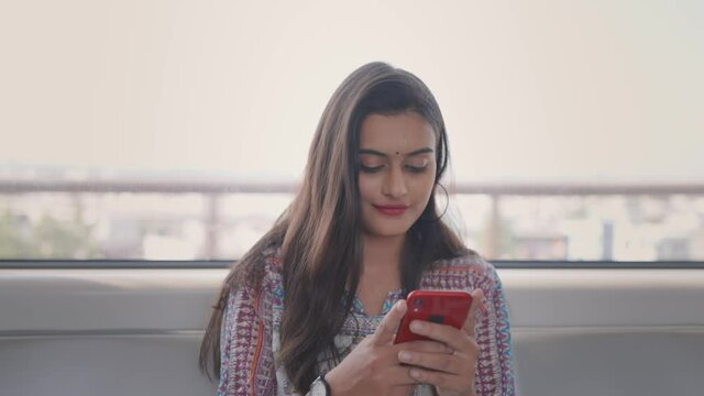 front view close shot of a young indian female sitting in a  moving city metro railway and using a mobile phone to type a text message with smile on face
