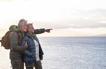 Two carefree senior people with backpack on their back enjoys the hike on the ocean cliffs looking at the horizon over water viewing the profile of an island and a ship. - Powered by Adobe