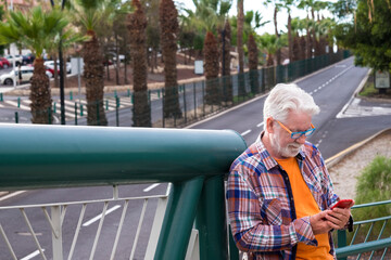 A white-haired senior man standing on an elevated city street with cellphone in the hand reading a message. Old people using modern technology. Boulevard behind him with palm trees