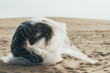 Female sorrow. Defocused silhouette. Isolation loneliness. Profile of depressed woman in black sitting on knees clasping head in polyethylene film flying in wind on sand desert skyline out of focus.