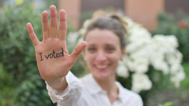Outdoors female portrait of young adult woman in white shirt showing her hand with slogan 'I voted' on flowers background, elections day concept