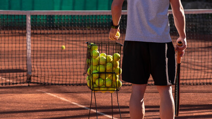 Tennis coach conducts training on red clay court, basket with tennis balls near him. Blurred background. Sports activity and leisure concept. Lifestyle - obrazy, fototapety, plakaty