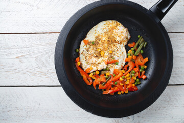 fried eggs and vegetables in a black frying pan on a light wooden table, close-up, space for text