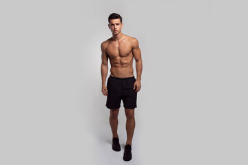 Full length image of a confident young man shirtless torso showing six pack abs, isolated grey background.