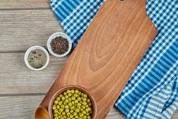 A bowl of boiled green peas with a spoon, spices, and a blue tablecloth on a wooden table