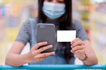 A masked woman is using a smartphone and in her hand is a white credit card. At the supermarket during the COVID-19 pandemic