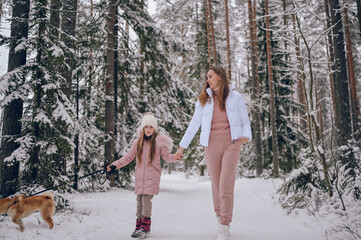 Happy family young mother and little cute girl in pink warm outwear walking having fun with red shiba inu dog in snowy white cold winter forest outdoors. Family sport vacation activities.