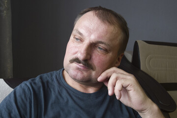 An elderly man with a mustache of Caucasian race in a gray T shirt in the interior is sitting thinking looking out the window