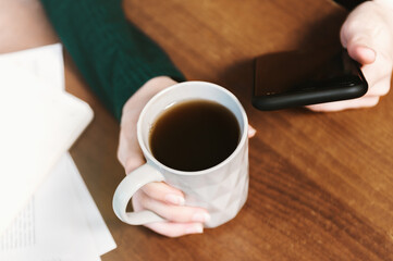 Female hands hold a gray mug with tea and write a message in the smartphone. Surfing the internet, lunch break at home office. Breakfast at the desk. Modern urban lifestyle