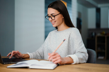 Happy young woman writing down notes while working with laptop