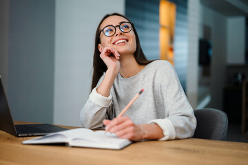 Happy young woman writing down notes while working with laptop