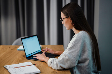 Brunette focused young woman in eyeglasses working with laptop at home