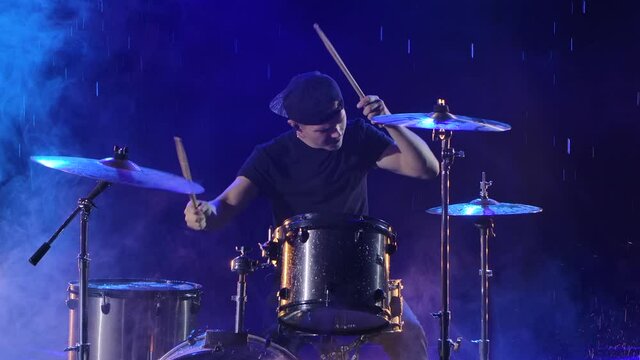 Young man enthusiastically plays the drum kit with drumsticks. A rock musician hits drums making music in the rain in a dark studio with blue lights. Close up. Slow motion.