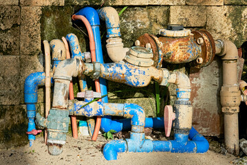 Confusing detail of rusty water meters, valves and blue pvc pipes outside of a dirty house wall