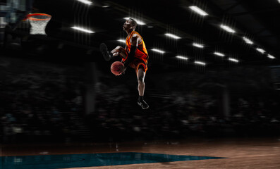 In high flight. African-american young basketball player in action and motion in flashlights over dark gym background. Concept of sport, movement, energy and dynamic, healthy lifestyle. Arena's