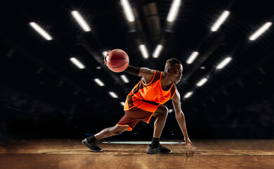 Team supporting. African-american young basketball player in action and motion in flashlights over dark gym background. Concept of sport, movement, energy and dynamic, healthy lifestyle. Arena's