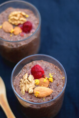 Two glasses of chocolate, orange and chia seed pudding with nut and raspberry topping. Selective focus, dark blue background.