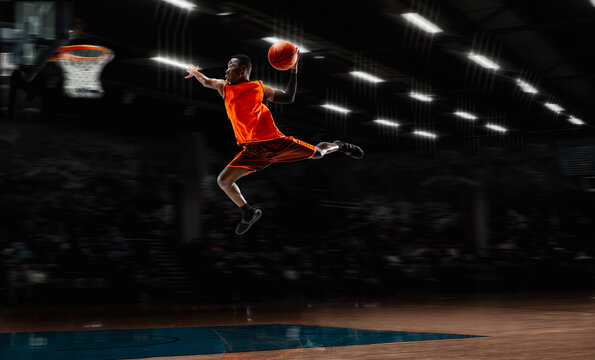 High dunk. African-american young basketball player in action and motion in flashlights over dark gym background. Concept of sport, movement, energy and dynamic, healthy lifestyle. Arena's drawned.