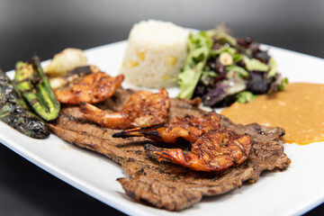 Grilled carne asada steak topped with split grilled shrimp served on a plate with rice and beans.
