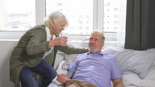 an elderly caring woman checks her husband's blood pressure and gives her sick husband a glass of water to drink at home on the bed. A woman's concern for a man.