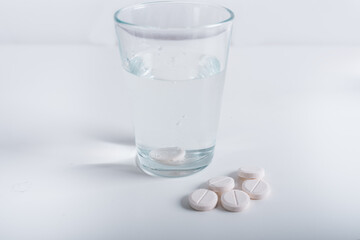 A glass of pure water on a white background, in which is a dissolving vitamin that forms bubbles. Next to it lie round pills. Disease treatment.