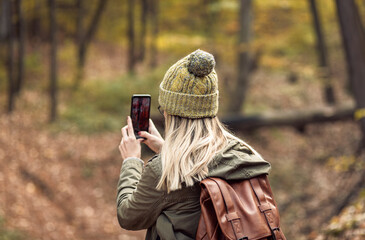 Young female hiker with backpack standing in forest and photographing nature with smartphone.