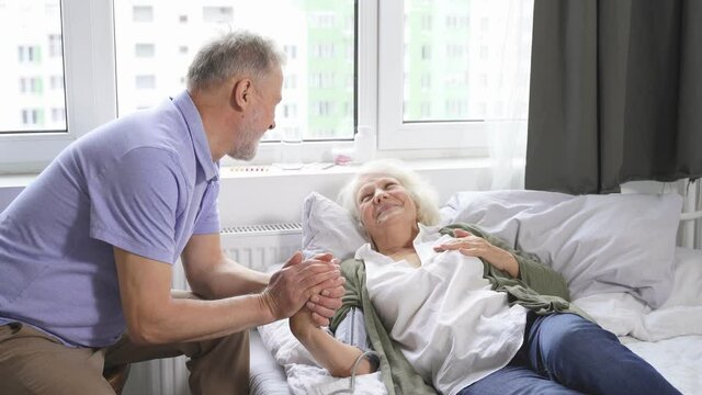 caring elderly man measures his beloved wife's blood pressure while lying on a bed in a room. The husband's concern for his wife.