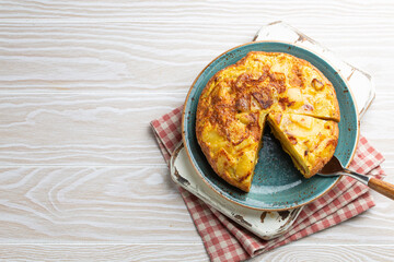 Homemade Spanish tortilla with one slice cut - omelette with potatoes on plate on white wooden rustic background top view. Traditional dish of Spain Tortilla de patatas for lunch or snack, overhead

