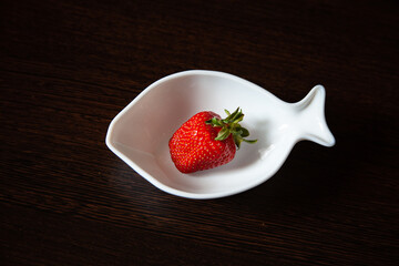 large horizontal photo. one big red strawberry in a white deep plate in the shape of a fish on a background of a dark wooden table. summer time
