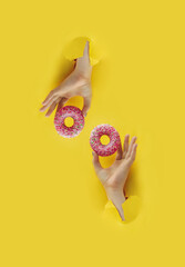 two hands holds delicious pink round glazed donut with sprinkle in torn yellow bright paper hole, vertical popular food blog content, selective focus