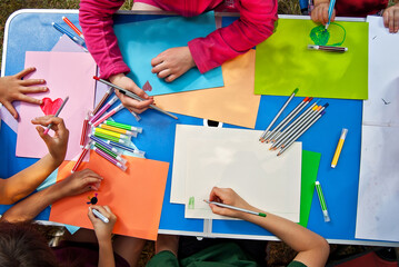 Children draw in nature. Lots of colored papers and pencils on a blue table. Boys and girls play outdoors. Drawing lesson in the park. Rest in a tent camp in the summer.